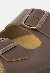 Sandale en cuir marron avec deux brides réglables, boucles en ton cuivre et design découpé. Semelle intérieure en suède beige doux pour un confort optimal.