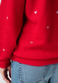 Pull rouge en tissu doux, orné d'un motif de petits cœurs argentés, avec des poignets et un ourlet côtelés. Associé à un jean bleu clair.