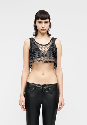 Jeune femme aux cheveux courts et foncés portant un crop top en maille noire par-dessus un soutien-gorge noir et un pantalon en cuir noir, sur un fond uni.