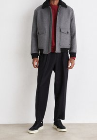 Man som bär en grå jacka med svart krage, röd tröja, svarta byxor och svarta sneakers med vita sulor som står mot en vit vägg.