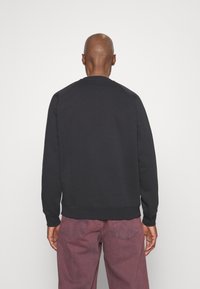 Man med rakad skalle som bär en enkel svart sweatshirt och urtvättade vinröda byxor, stående vänd mot en vit vägg med händerna vid sidorna.