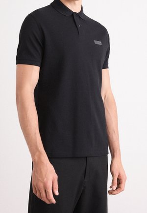 Mężczyzna noszący czarną koszulkę polo z krótkim rękawem i małym logo Barbour International na piersi.