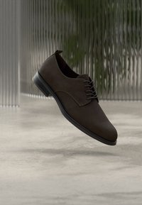 Καφέ σουέντ παπούτσι με κορδόνια, με λείο φινίρισμα, στρογγυλερή μύτη και μαύρη καουτσούκ σόλα. Μινιμαλιστικός σχεδιασμός με κορδόνια και ραφές στο ίδιο χρώμα.