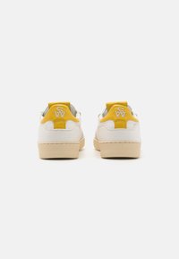 Baskets blanches avec un talon jaune et un logo. Cuir texturé et un embout lisse. Semelle extérieure crème et détails de couture minimalistes.