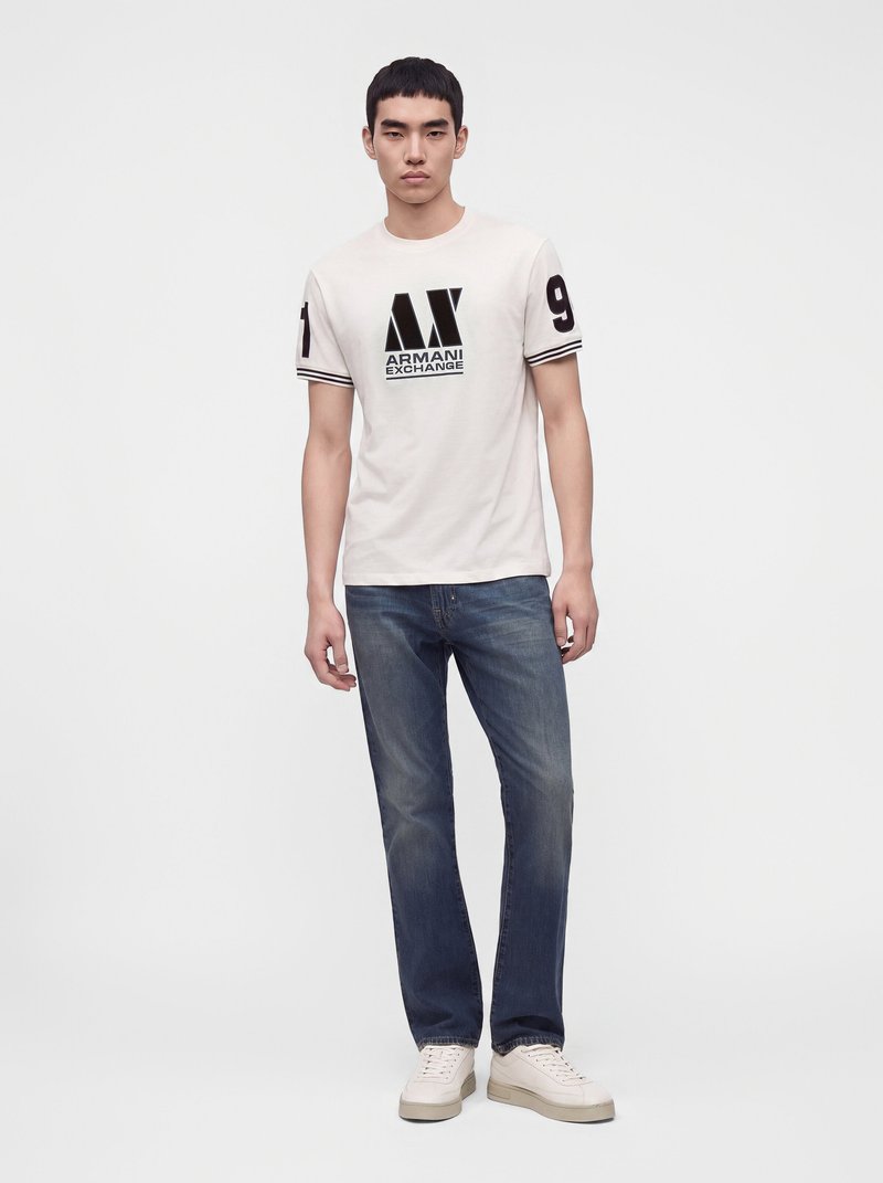 Млад мъж, облечен с бяла тениска Armani Exchange с черен принт и сини дънки на едноцветен бял фон.