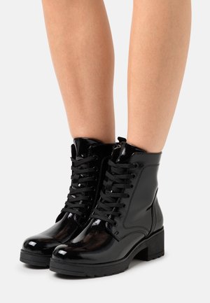 Bottines noires brillantes en cuir verni à lacets avec semelles épaisses portées sur des jambes nues sur un fond clair uni.