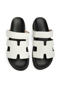 White vegan leather slide sandals with two wide straps, a distinctive cut-out design, and a black textured sole, featuring brand logo.