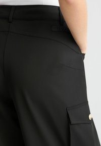 Zwarte broek van glad materiaal, met een achterzak met een gouden knoopaccent en een label met de tekst "LIUJO."