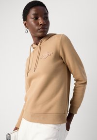 Felpa con cappuccio beige con una finitura testurizzata, caratterizzata da un logo frontale e coulisse. Maniche lunghe, polsini a coste e dettaglio all'orlo.