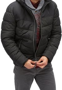 Giacca a vento nera con cappuccio, frontale con zip e cuciture orizzontali. Indossata sopra un maglione grigio e rosso a fantasia. Maniche con polsini regolabili.