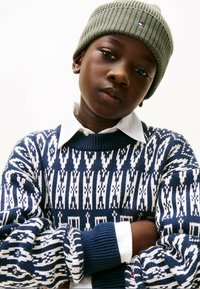 Navy gestrickter Pullover mit weißem geometrischen Muster, über einem weißen Hemd mit Kragen getragen. Olivgrüne gerippte Mütze mit Logo.