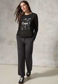 Schwarzes Langarmshirt mit silbernem grafischem Text, kombiniert mit lockeren schwarzen Hosen und schwarzen Sneakers, vor einem neutralen Hintergrund.