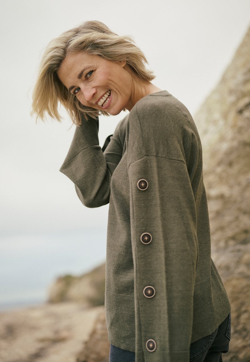 Suéter de punto verde oliva con un ajuste relajado, que presenta un cuello en V, mangas largas y grandes botones a lo largo del brazo izquierdo. Tela con textura.