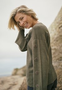 Pull tricoté vert olive à coupe décontractée, avec un col en V, des manches longues et de gros boutons le long du bras gauche. Tissu texturé.