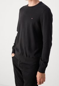 Felpa nera in cotone con collo a giro, maniche lunghe, polsini e orlo a costine, con un piccolo logo sul petto. Indossata con pantaloni neri.