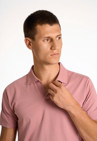 Ljusrosa polo skjorta med klassisk krage, korta ärmar och en texturerad tyg. Modellen justerar kragen med ena handen.