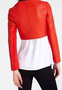 Rode cropped jas met ribbeltextuur, voorzien van lange mouwen en een ronde halslijn. G gedragen over een wit topje en zwarte broek.