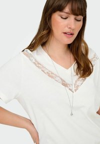 Bílý bavlněný T-shirt s výstřihem do V, zdobený průhlednou krajkovou vsadkou. Krátké rukávy, hladká textura, v kombinaci se stříbrným náhrdelníkem a přívěskem.