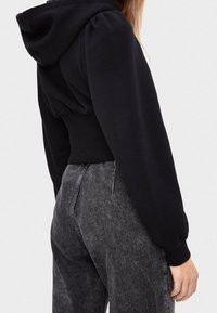 Felpa corta nera con maniche a sbuffo e fascia in vita abbinata, realizzata in materiale morbido. Abbinata a jeans grigi a vita alta.