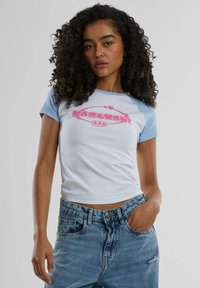 Camiseta blanca con mangas raglán azul claro, que presenta un logo rosa de "Karl Kani" en la parte delantera. El material parece suave, diseño corto.