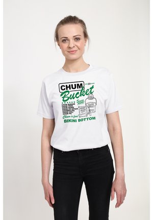 Vrouw draagt wit T-shirt met opdruk "Chum Bucket Bikini Bottom" waarop menu-items staan, staand tegen een effen lichte achtergrond.