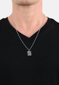 Collana con pendente in argento con un'etichetta rettangolare caratterizzata da testo inciso, appesa a una sottile catena, indossata su una maglietta nera a scollo a v.
