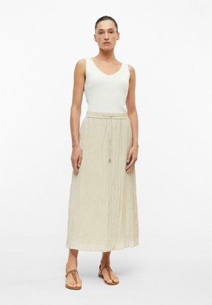 Femme debout portant un haut blanc sans manches, une jupe midi beige à cordon de serrage et des sandales plates marron sur un fond blanc uni.