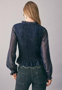 Mornarska bluza z teksturiranim, gubanim hrbtom, prosojnimi dolgimi rokavi in volanastim ovratnikom; v kombi z črnimi kavbojkami z okrasnimi studenčki.