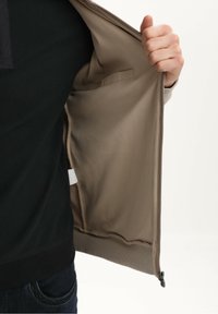 Beżowa kurtka rozpięta, odsłaniająca gładką, miękką podszewkę z boczną kieszenią, prezentująca bezszwowy design oraz detale zamka na dole.