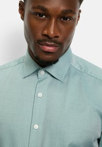 Hellgrünes Hemd mit durchgehender Knopfleiste und strukturiertem Karomuster. Verfügt über einen klassischen Kragen und weiße Knöpfe, die das dezente Design des Stoffes hervorheben.