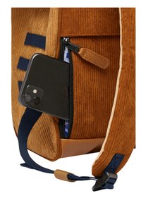 Zaino in velluto a coste marrone con accenti in pelle, dotato di una tasca con zip che contiene uno smartphone nero. Spallacci blu visibili e rivestimento interno.