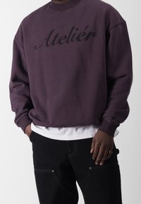 Lilla sweatshirt med broderet "Ateliér" tekst i sort, rund halsudskæring og løs pasform, parret med sorte bukser og en hvid undertrøje.
