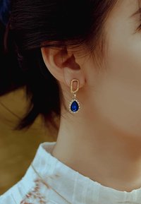 Boucles d'oreilles en or en forme de créole avec une pierre précieuse bleue en forme de poire, entourée de cristaux transparents. Texture lisse avec des détails complexes.