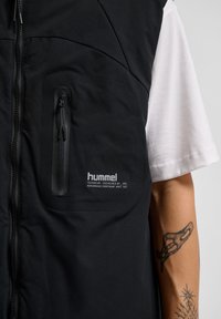 Schwarze technische Weste mit einer Reißverschluss-Brusttasche, glatter Textur und Logodetail auf der unteren rechten Seite. Weißes Shirt darunter sichtbar.
