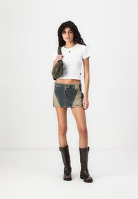 T-shirt bianca corta, minigonna in denim con pannelli verdi e beige, e stivali al ginocchio marroni con tacco, abbinati a una borsa verde.