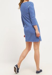 Robe bleu et blanche à rayures, longueur genou, avec des manches trois-quarts et des fentes sur les côtés. Portée avec des baskets sombres sur un fond clair.