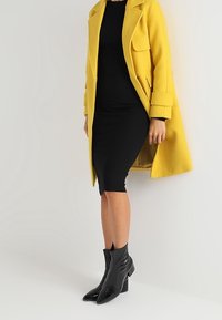 Gelber Wollmantel mit übergroßen Revers, zwei vorderen Taschen, kombiniert mit einem schwarzen gerippten Kleid und glänzenden schwarzen Stiefeletten mit spitzen Zehen.