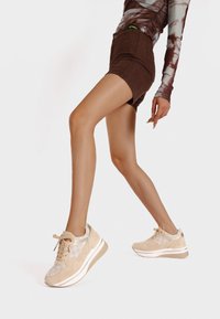 Sneaker beige con un motivo testurizzato e lacci, caratterizzati da una suola più spessa e sottili accenti bianchi. Indossati con pantaloni corti marroni e una maglietta fantasia.