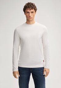 Weißes langärmeliges T-Shirt aus weichem Stoff, mit Rundhalsausschnitt und schmaler Passform, kombiniert mit dunkelblauen Jeans. Sichtbares rotes Etikett.