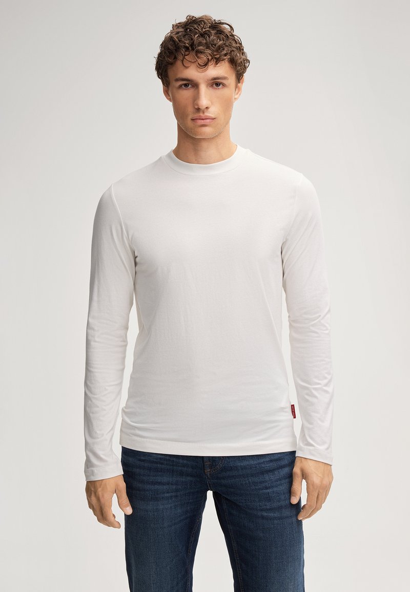 Weißes langärmeliges T-Shirt aus weichem Stoff, mit Rundhalsausschnitt und schmaler Passform, kombiniert mit dunkelblauen Jeans. Sichtbares rotes Etikett.