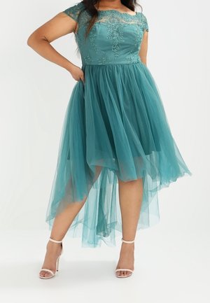 Femme portant une robe haute-basse en dentelle et tulle bleu sarcelle avec des manches courtes, associée à des escarpins blancs à bride à la cheville, debout avec une main sur la hanche.
