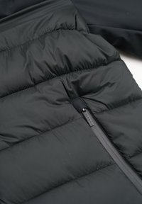 Schwarze Daunenjacke mit einer glatten, glänzenden Oberfläche, die über gestepptes Muster und eine seitliche Reißverschlusstasche mit matter Oberfläche verfügt.