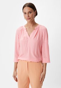 Rosa Bluse mit kurzen Ärmeln, V-Ausschnitt und gerafft verarbeitetem Halsrand. Weicher Stoff mit lockerer Passform, kombiniert mit hellpfirsichfarbenen Hosen.