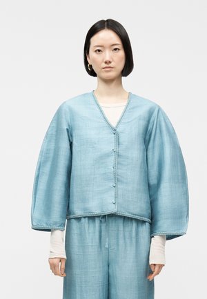 Kvinde med glat sort hår iført lyseblå, tekstureret, oversize jakke og matchende vidde bukser over en hvid langærmet top.