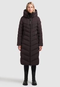Vrouw in een lange zwarte gewatteerde pufferjas met capuchon, ritsluiting en met rits afgesloten zakken, gecombineerd met zwarte leggings en zwarte laarzen.