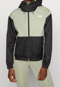 Wiatrochron w jasnozielonym i czarnym kolorze, z zamkiem z przodu, regulowanym kapturem, elastycznymi mankietami, bocznymi kieszeniami oraz akcentami logo.