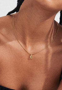 Goldene Halskette mit feiner Kette und einem kleinen, runden "C"-Anhänger. Der Hintergrund zeigt Hauttöne und eine einfache, neutrale Textur.