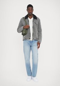 Veste en velours côtelé grise avec un col noir, chemise blanche, jeans bleu clair et baskets blanches. Porte un petit sac bandoulière vert.
