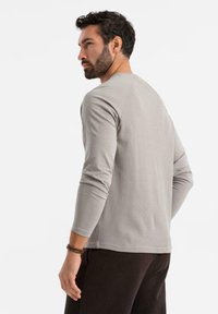 Camisa de manga larga gris claro con textura suave y acanalada; diseño ajustado con cuello redondo y mangas ligeramente ajustadas.