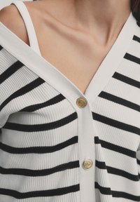 Gros plan d'une personne portant un cardigan blanc avec des rayures horizontales noires et des boutons dorés, montrant une bretelle blanche en dessous au niveau de l'épaule.